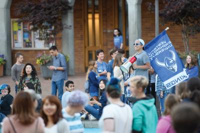 A student stands on the edge of Fisher Fountain with a bull horn and a WWU Viking blue and white flag and is surrounded by other students in Red Square