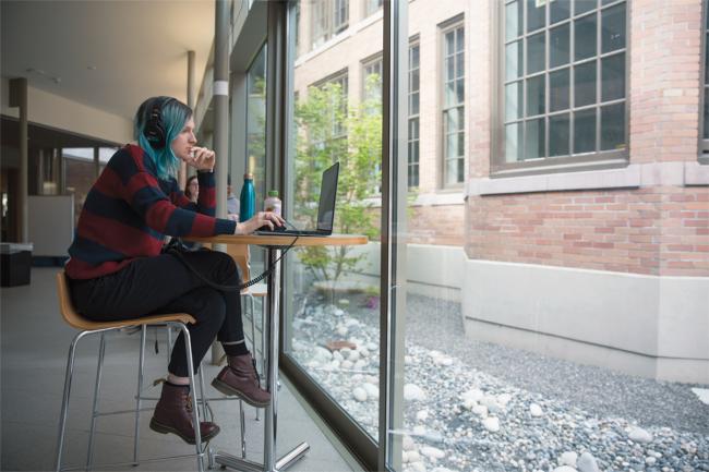 Female student with blue hair working on her laptop at a table in the Miller Hall Collaborative Space 