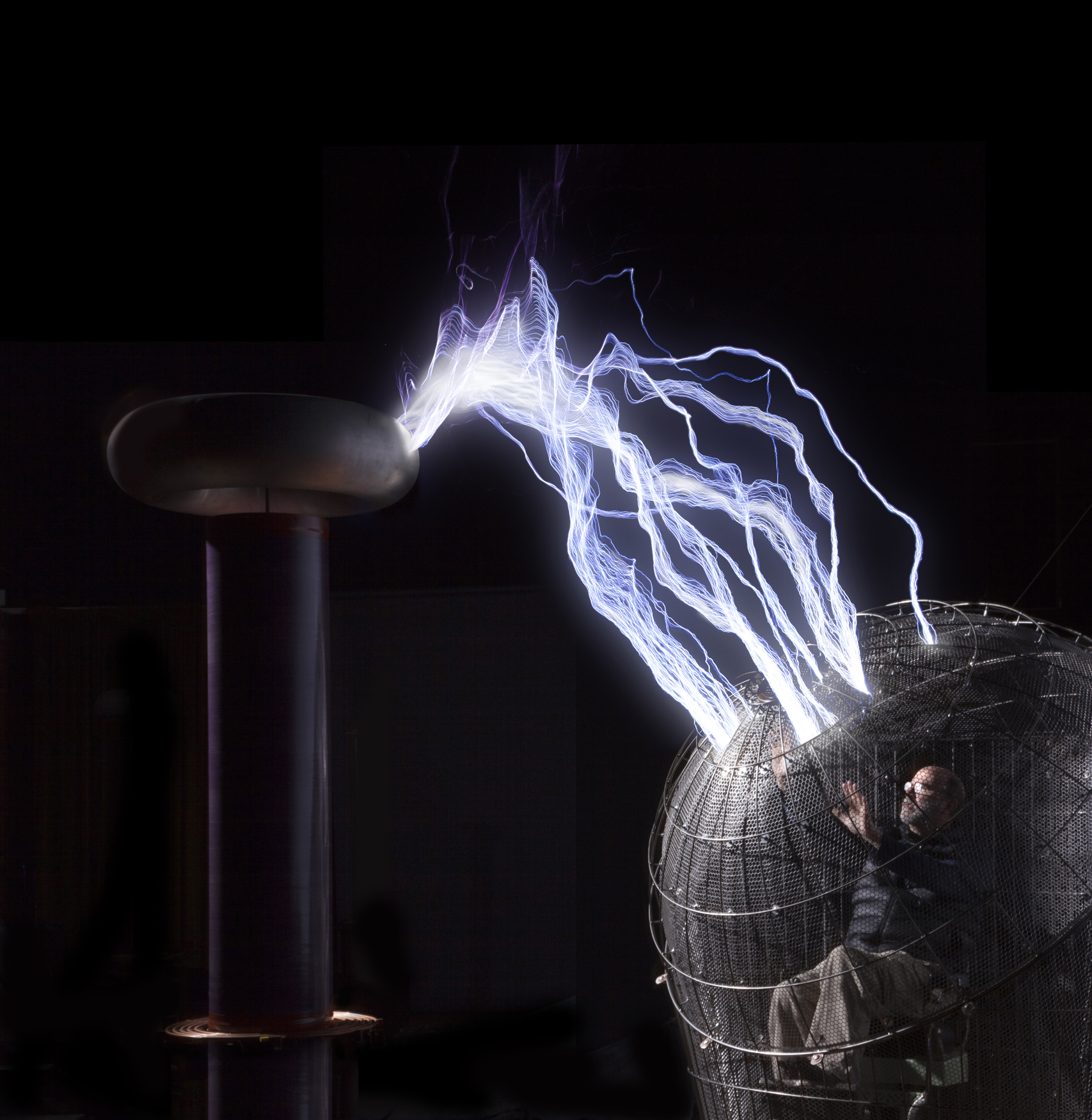 man crouched in a metal cage shaped like a ball with lightning bolts shooting at from a tall metal rod.